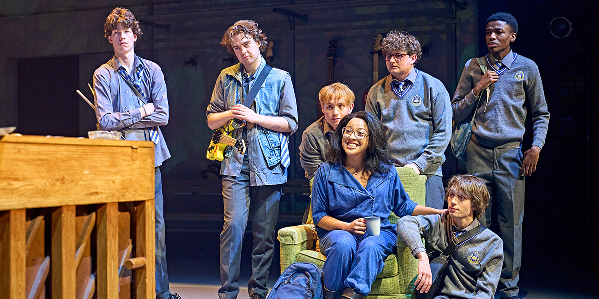 A group of six students in school uniforms on a stage set, surrounding a person sitting in a green armchair.