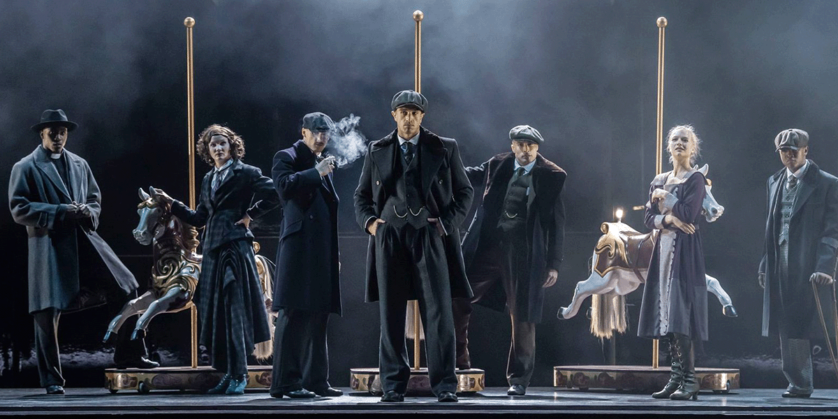 A dancer in a flat cap performs on stage, surrounded by dramatic lighting and dark, textured backdrops.
