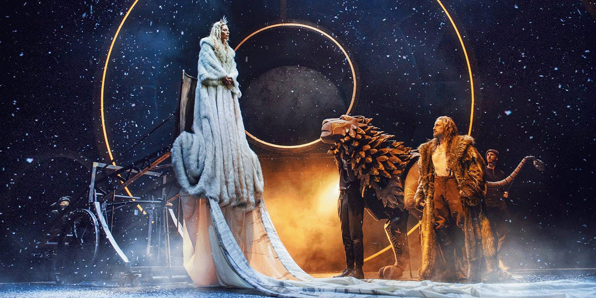 Three performers stand against a vast starry sky. One is dressed in white furs, another is handling a life-sized wooden lion puppet, and the third is wearing brown furs.