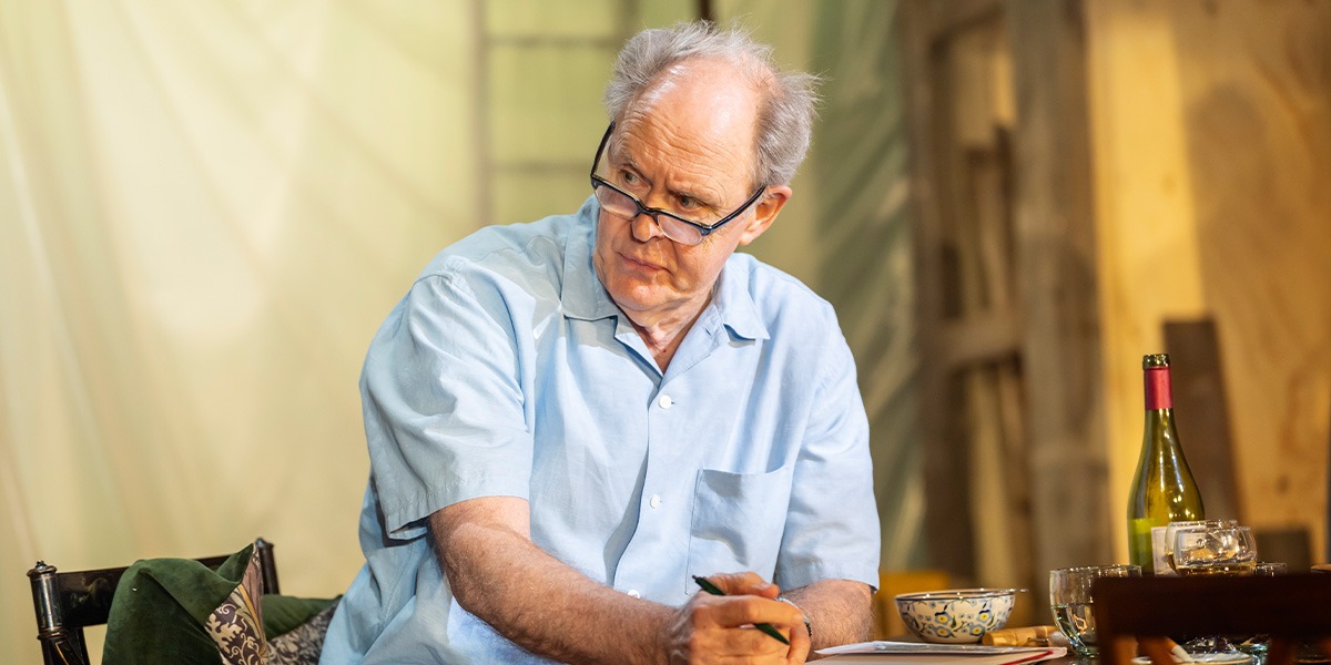 John Lithgow (Roald Dahl) is wearing a pale blue shirt and reading glasses while sitting at a table writing.