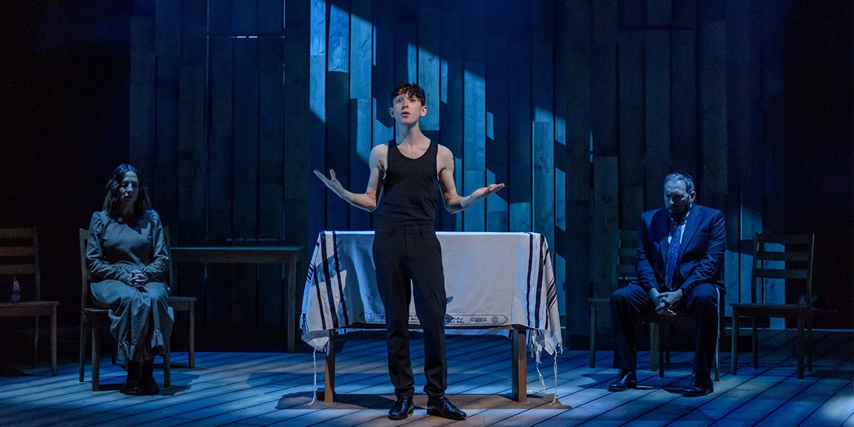 A person wearing a black vest and trousers is standing central holding both arms out on either side as if to ask a question.