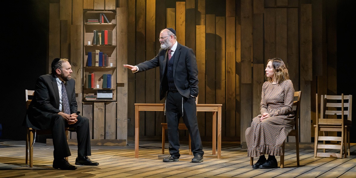 Two people are sitting on chairs on either side. In the centre a person wearing a black suit is standing holding out their hand as if to make a 'stop' gesture.