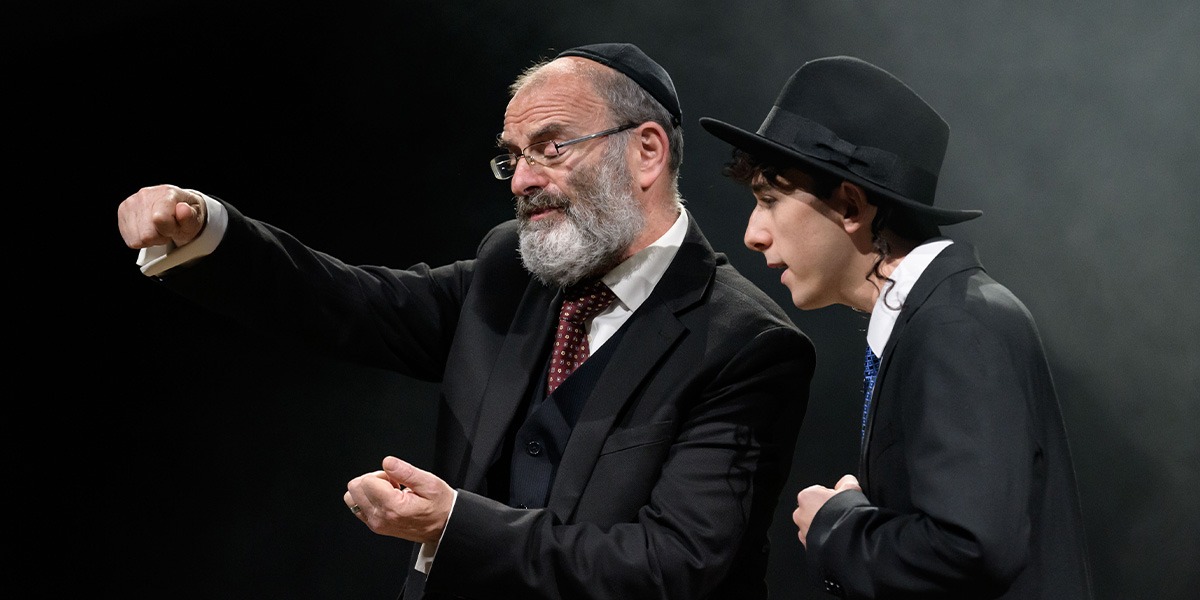 Two people wearing black suits are standing together. They are both looking down as if reading something.