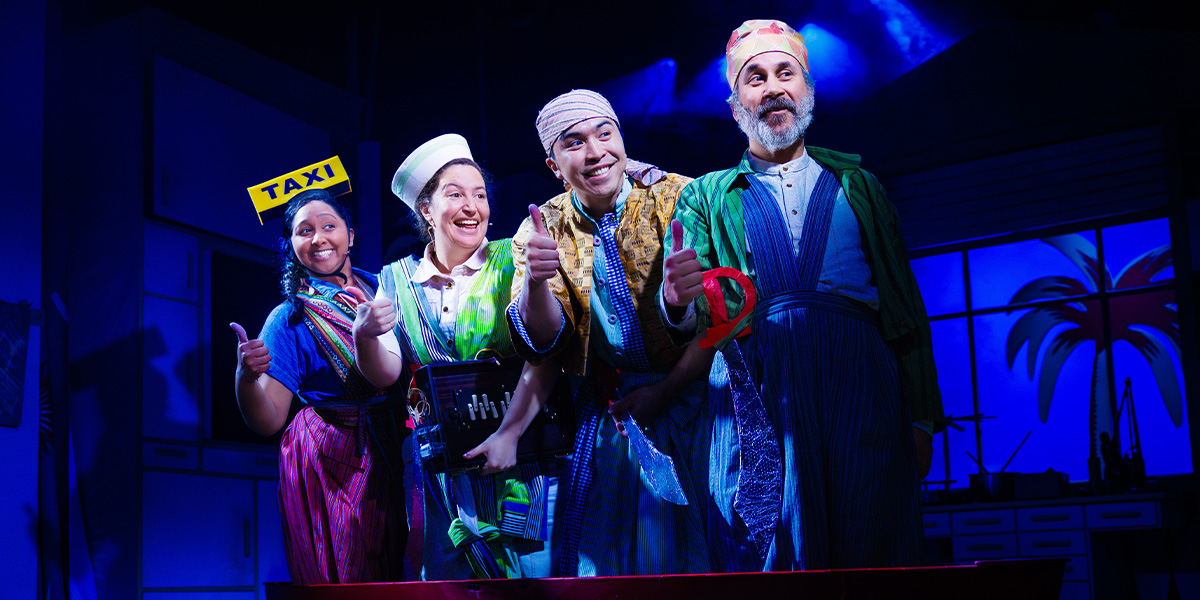 Four people in vibrant costumes stand on stage bathed in warm purple light.