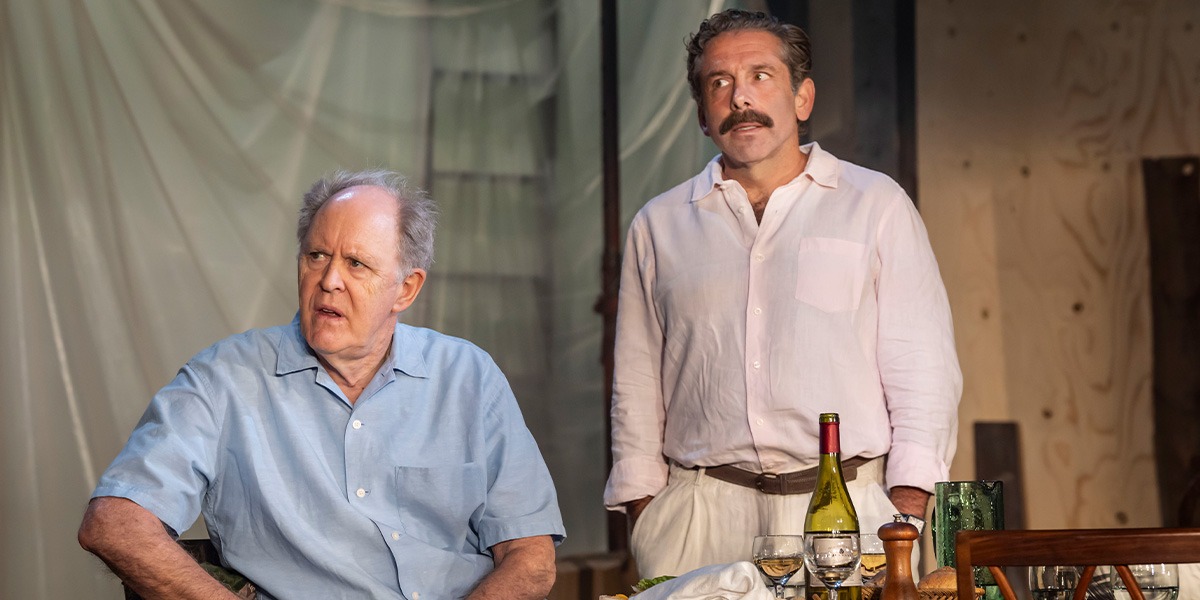 Elliot Levey (Tom Maschler) is standing beside John Lithgow (Roald Dahl) who is sitting at a table.