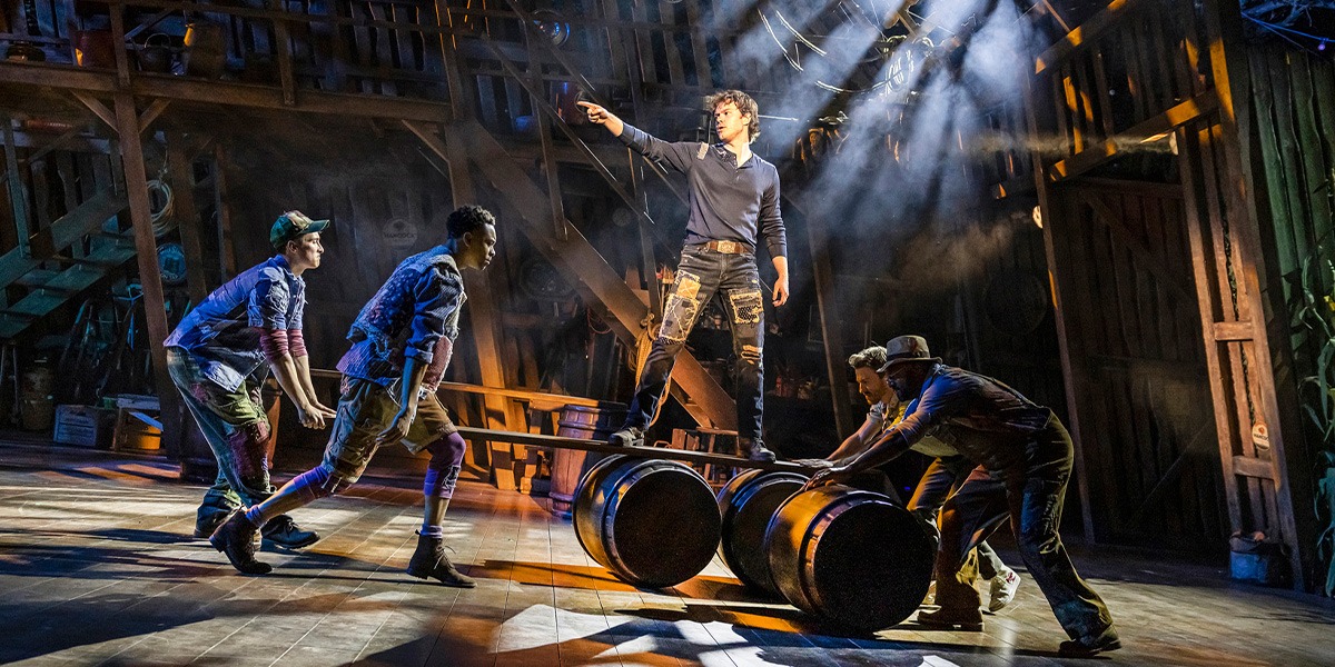 A dynamic scene from Shucked featuring a performer in the centre pointing upward, surrounded by others interacting with barrels on a wooden stage set, enhanced by dramatic lighting.