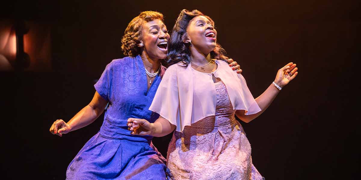 Beverley Knight and Ntombizodwa Ndlovu perform joyfully, wearing elegant dresses, against a warm, dimly lit background.