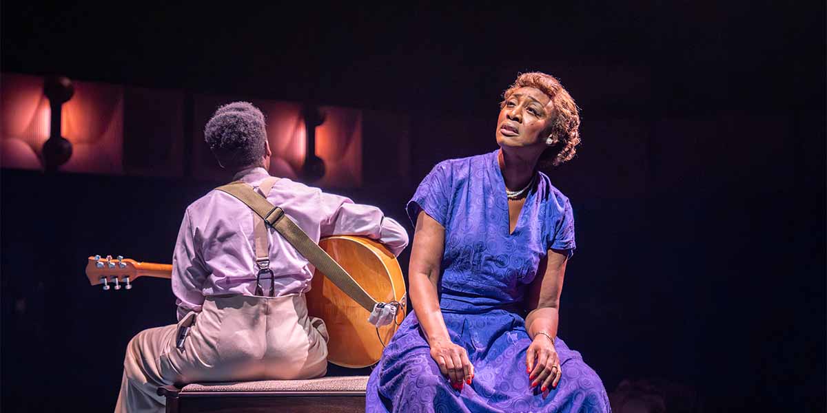 Beverley Knight as Sister Rosetta Tharpe, wearing a blue dress, gazes thoughtfully while seated beside a guitarist.