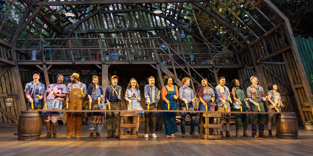 The image shows a dynamic stage scene from the musical Shucked with a group of performers in various costumes standing in a line.