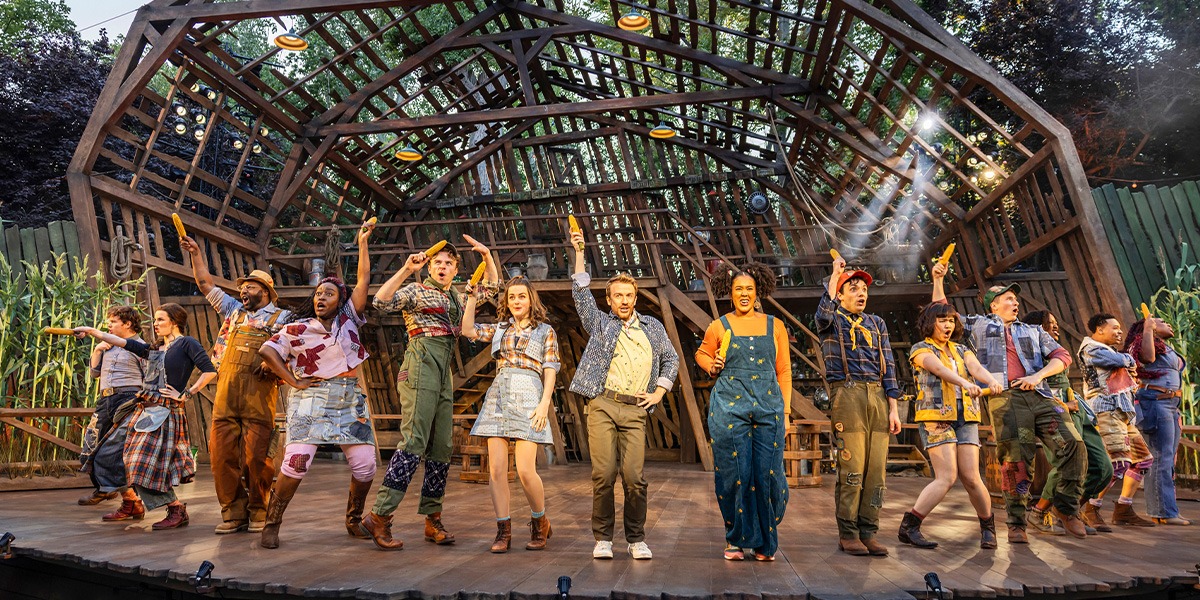 The image shows a dynamic stage scene from the musical Shucked with a group of performers in various costumes singing and dancing enthusiastically.