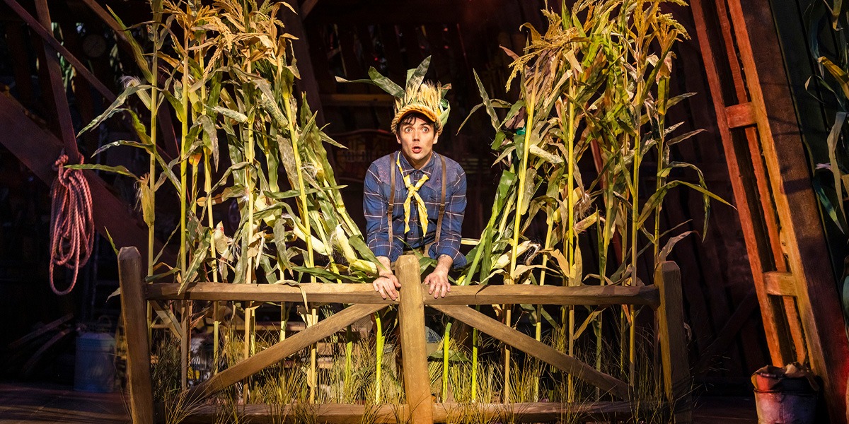 Keith Ramsay (Peanut) is sitting on a wooden fence surrounded by cornstalks on stage, wearing a straw hat and rustic outfit.