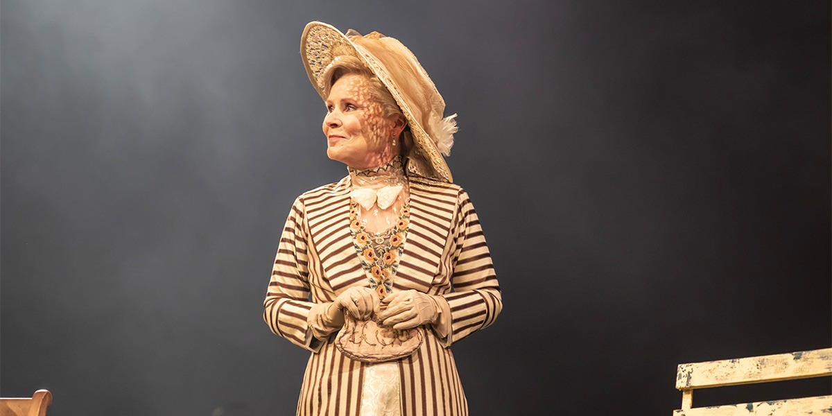 Imelda Staunton (Mrs. Kitty Warren) is wearing elegant brown and white striped attire and is looking towards the left smiling.