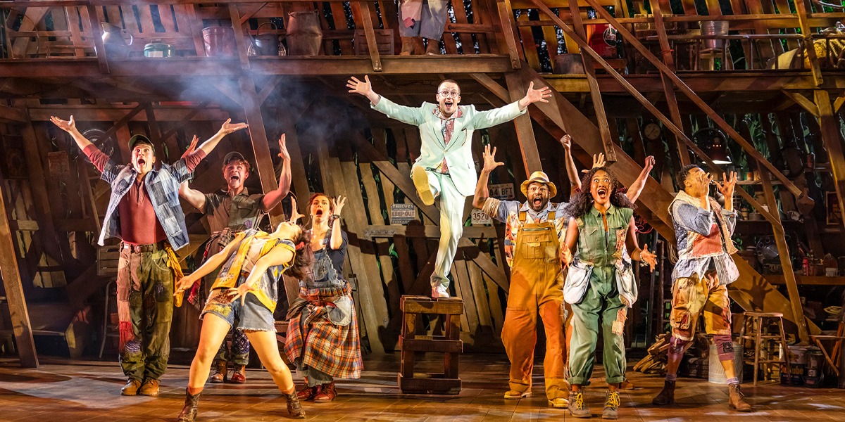 The image shows a dynamic stage scene from the musical Shucked with a group of performers in various costumes singing and dancing enthusiastically. One performer in the centre appears particularly exuberant, arms spread wide and beaming with joy.