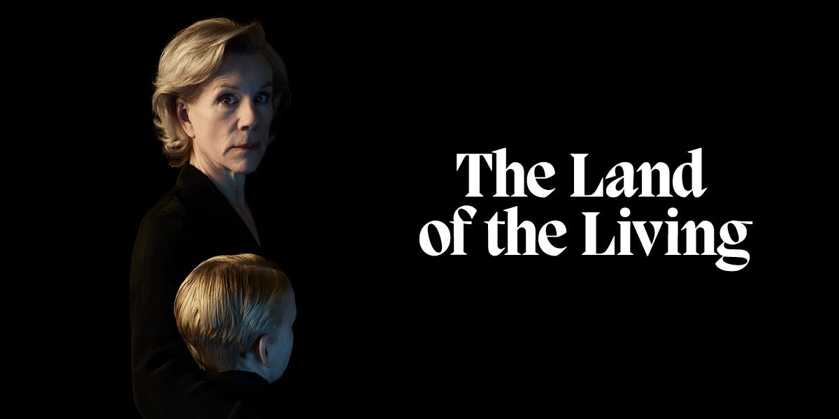 The background is black. On the right it reads ' The Land Of The Living' in white letters. On the left is an image of a person with shoulder-length, blonde hair. They are wearing a black jacket. In front of them is a child facing away from the camera. The child has short blonde hair and is also wearing a black jacket.