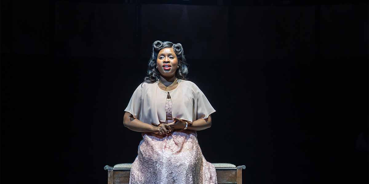 Ntombizodwa Ndlovu, dressed in a pale pink gown, performs passionately on stage, illuminated against a dark background.