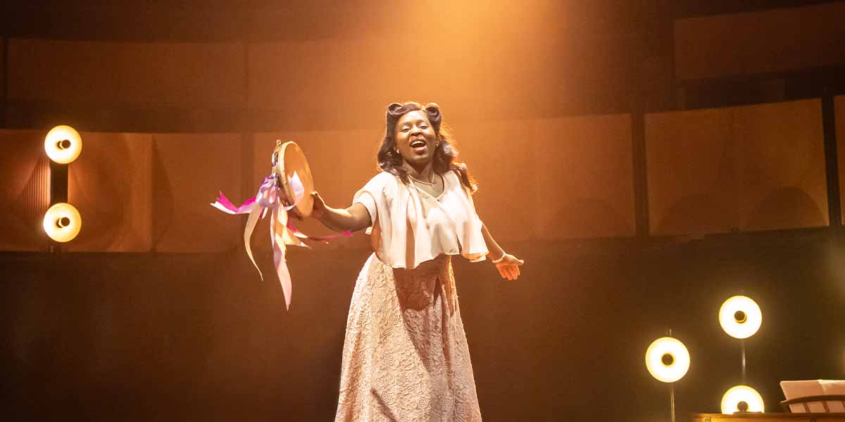 Ntombizodwa Ndlovu, portraying Marie Knight, joyfully performs with a tambourine on stage, illuminated by warm lighting.