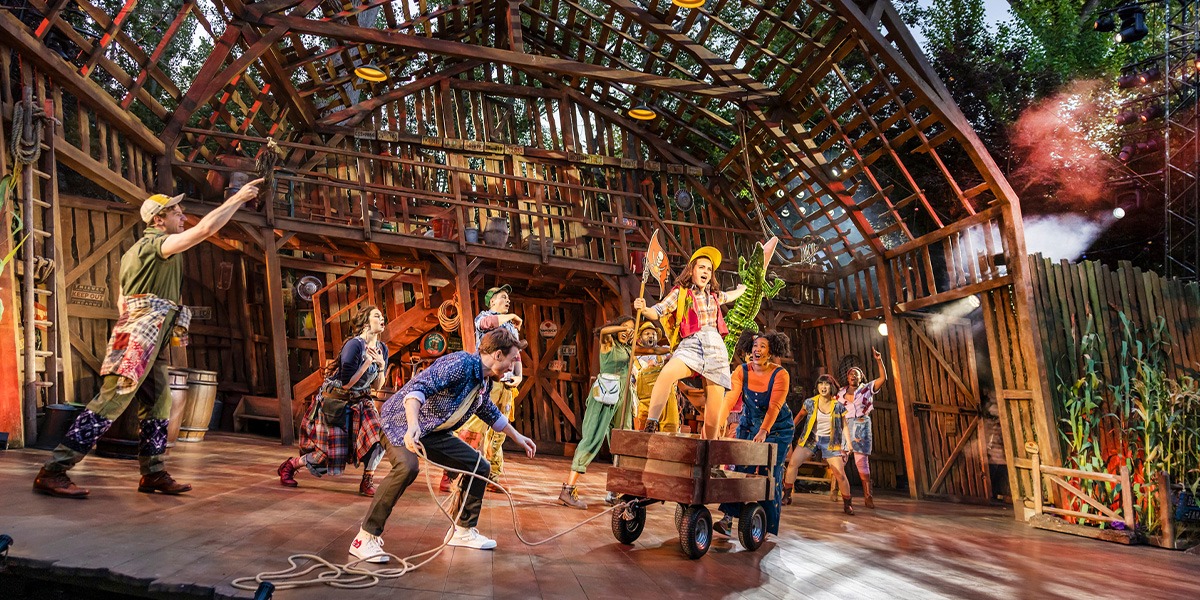 The image shows a dynamic stage scene from the musical Shucked featuring performers energetically dancing and interacting in a rustic barn set, with vibrant lighting and colorful theatrical effects.
