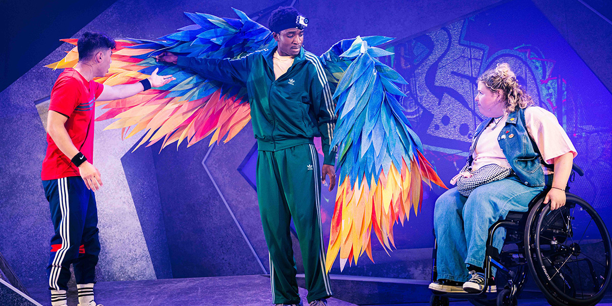Three individuals on a stage with a backdrop featuring mystical symbols; one person in a green tracksuit and large blue wings stands center, another to the left in a red and black sporty outfit touches one wing, and a third seated in a wheelchair with similar styled wings to the right interacts with the central figure.