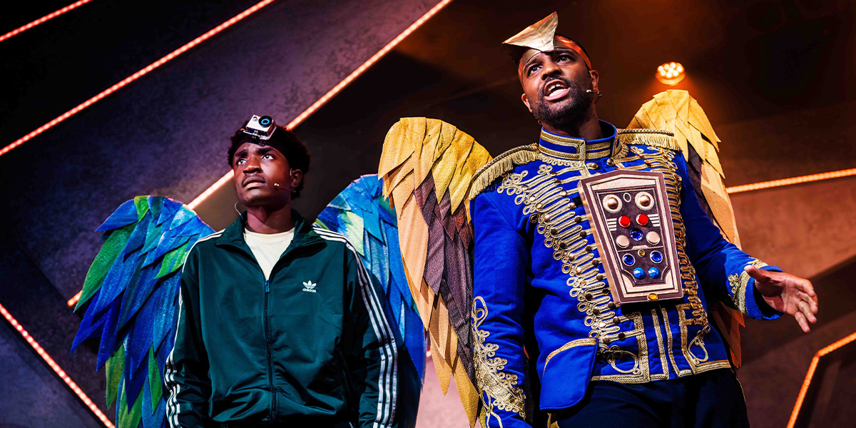 Two performers on stage, one wearing a casual outfit with a cap, and the other in an elaborate costume with large, colorful wings, set against a vibrant, geometrically patterned background.