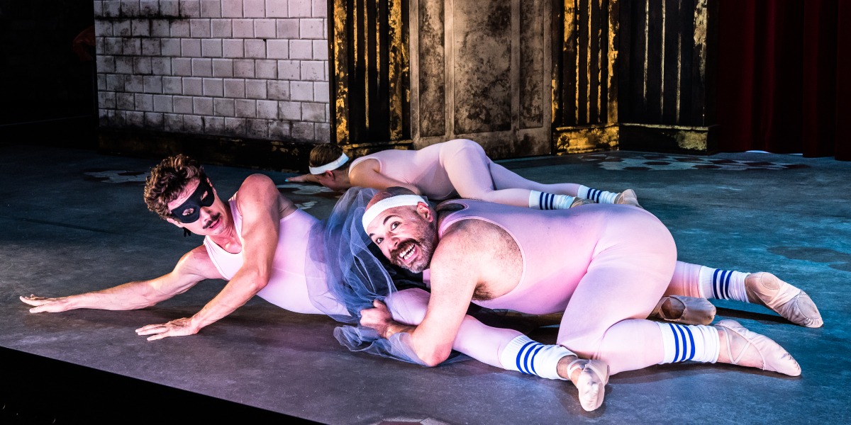 Three ballet dancers wearing pink leotards and headbands are humorously tangled on stage. One wears a black eye mask, and all have striped socks. The scene is lively, set against a backdrop of tiled and textured walls.