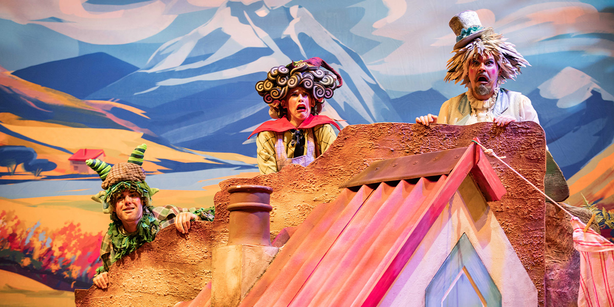 Performers in colorful costumes on a whimsically designed stage set with mountain backdrops, part of a theatrical production.