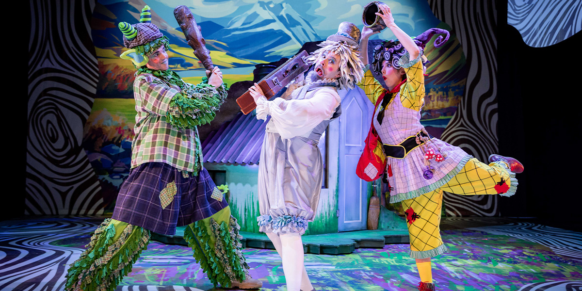 Three performers in colourful, whimsical costumes on stage, acting out a scene with exaggerated expressions and dynamic poses. One holds a guitar, surrounded by a vibrant set reminiscent of a fantastical landscape.