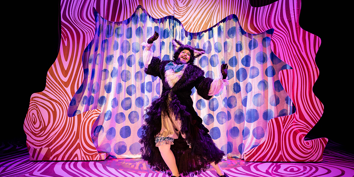 Performer in an elaborate costume dancing joyously on stage with a vibrant, swirling pink and blue backdrop.