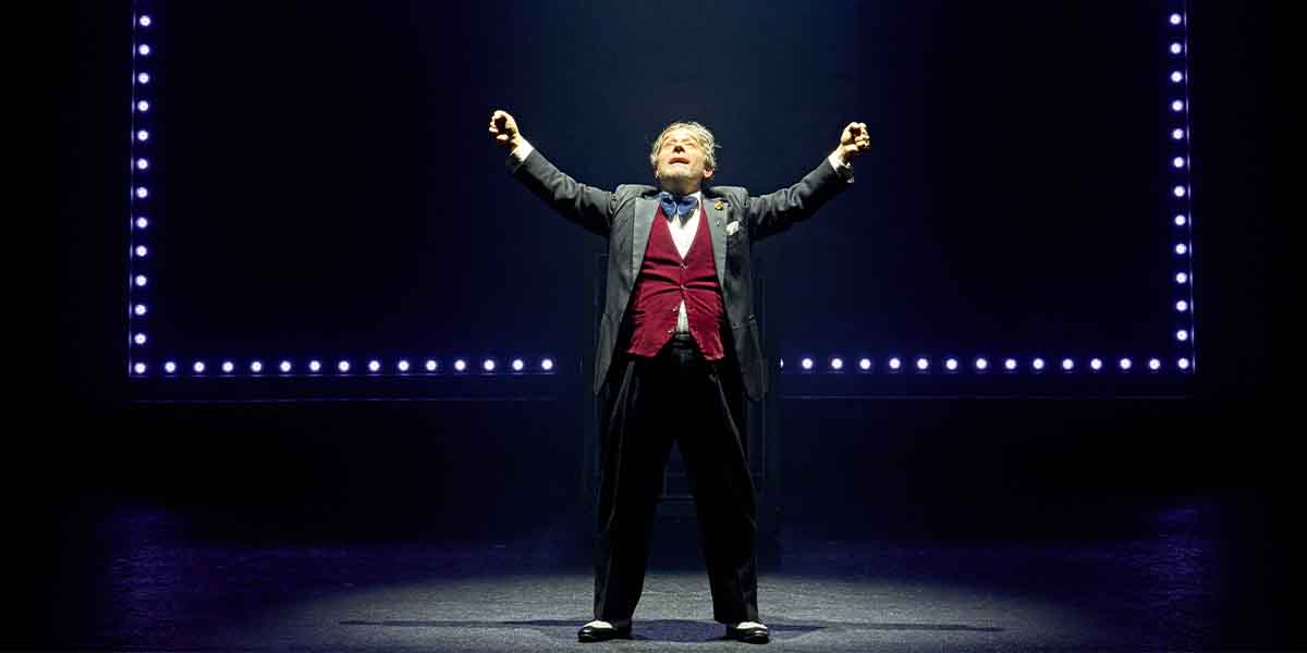 Andy Nyman as Max Bialystock, arms raised, in a theatrical pose against a dark background with lights.