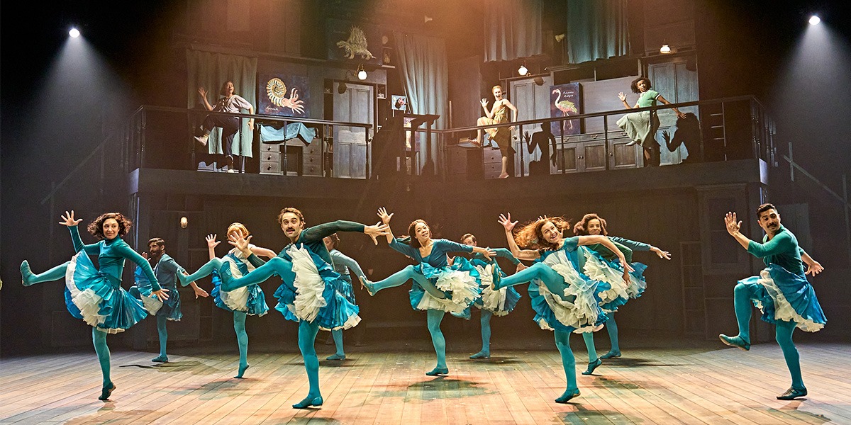 Dancers in turquoise costumes with white frills perform energetically on a wooden stage.
