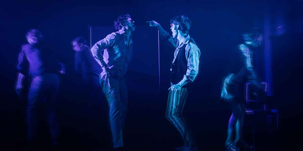 Calam Lynch and Noah Ritter perform energetically under blue lighting, surrounded by blurred figures in the background.