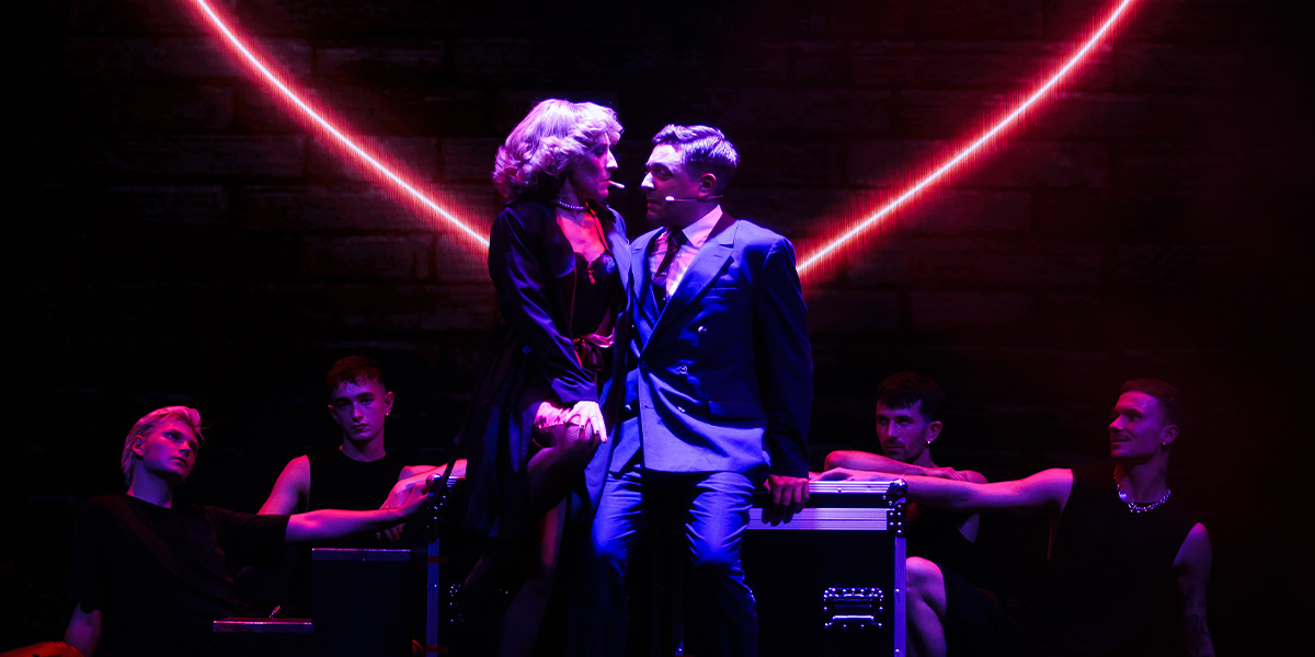 Two performers in an emotional scene on stage under red stage lights, with background actors observing.