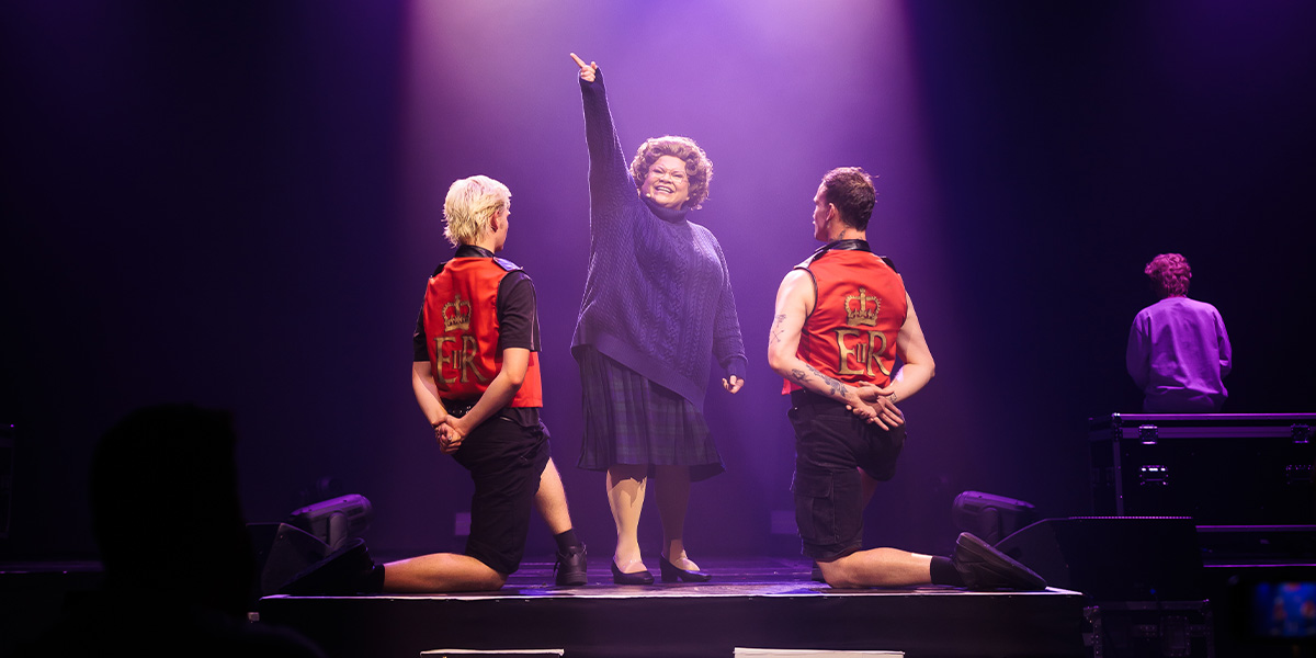 A person smiling broadly stands center stage, pointing upwards, while flanked by two individuals in red vests decorated with letters, all illuminated by dramatic stage lighting with a soft purple hue. They are participating in a theatrical performance.