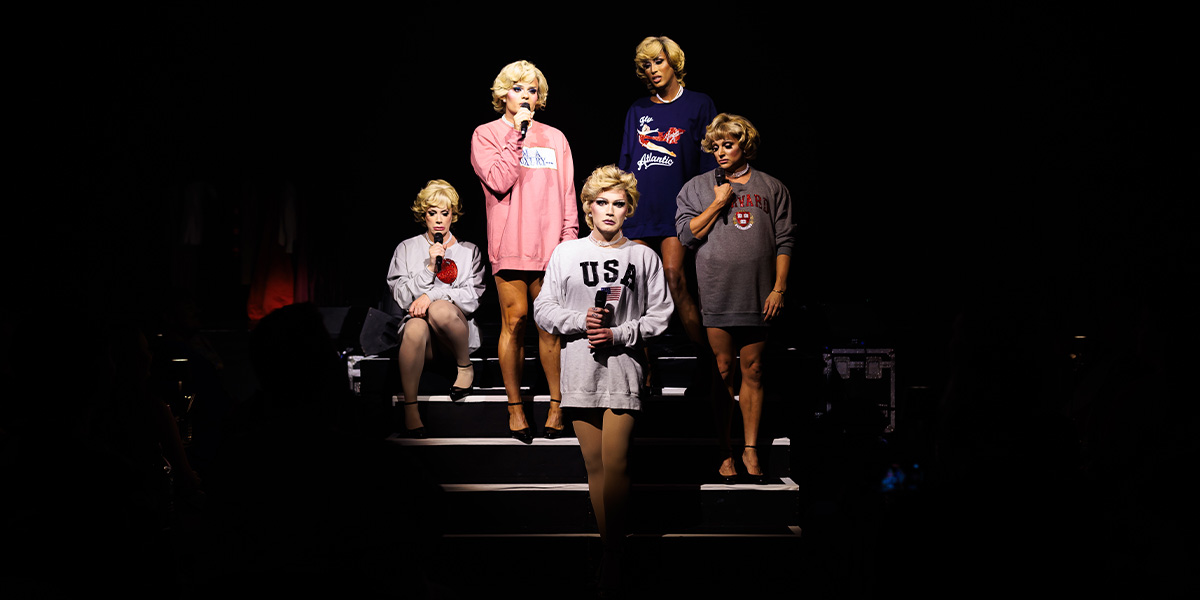 Five performers on a dark stage, wearing sporty attire including one in a "USA" sweater, during a theatrical show.