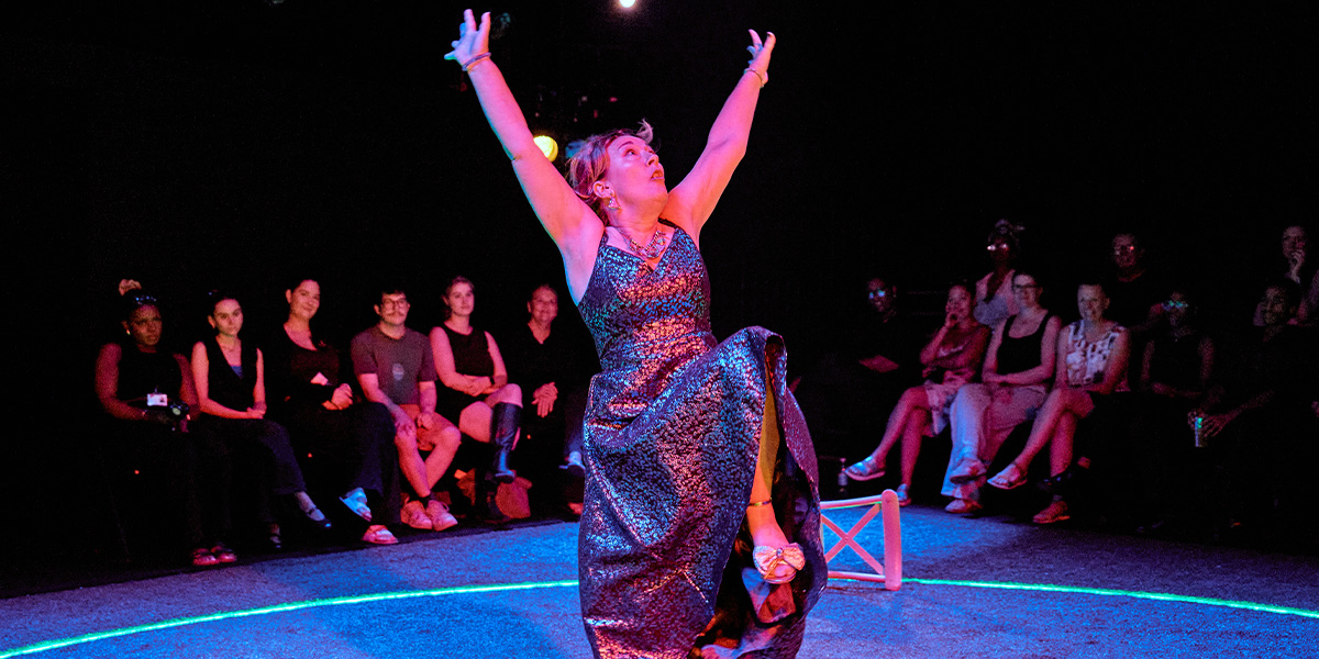 Performer in a glittery dress enthusiastically reaches upwards on stage during a live performance, with an attentive audience seated around in a dimly lit room.