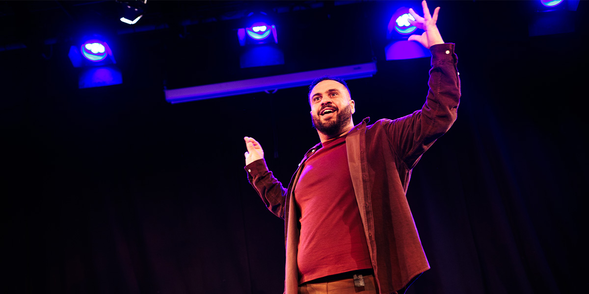 A performer gestures expressively on stage, wearing a maroon shirt and brown jacket, under blue lights.