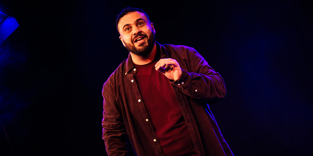 A man with a beard gestures while speaking, wearing a dark shirt over a maroon top, against a dark background.