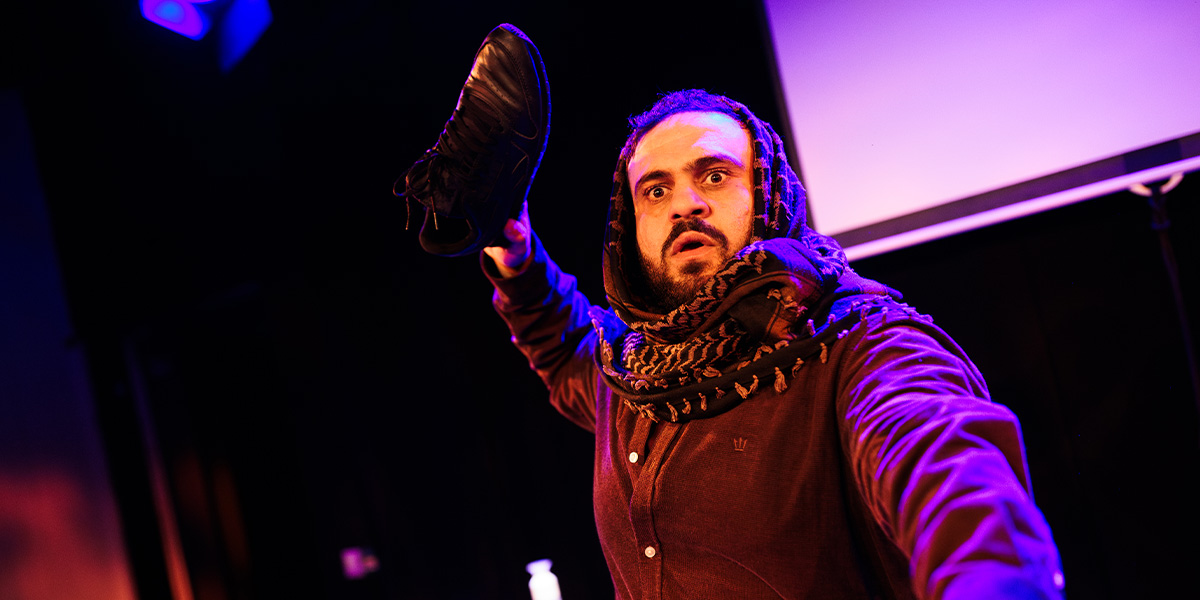 A performer with a scarf raises a black shoe, expressing surprise, against a dark background with purple lighting.