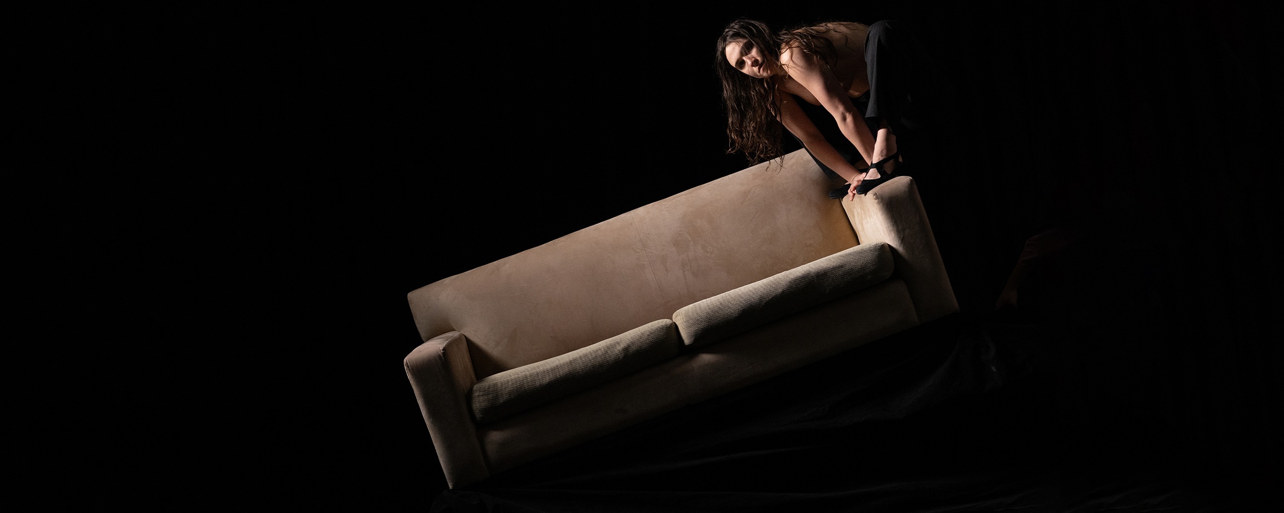 There is a black background. There is a cream coloured sofa, that is titlted half way into the air. On the right arm of the sofa is a woman with dark hair, half climbing on the arm. Her costume is obscured by the black background. One of her feet is visible with a black shoe.