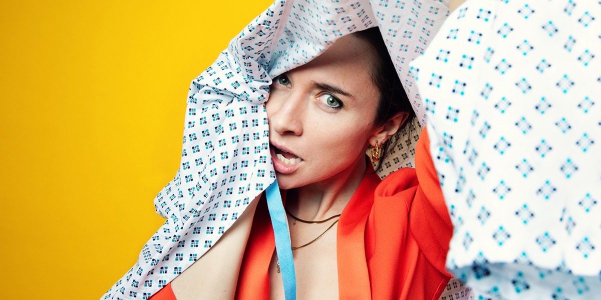 There is a yellow background. In the middle of the image is a woman wearing an orange top. She has a white hospital gown over her head, covering a part of her face.