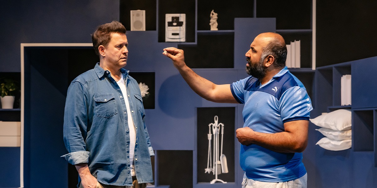 Two actors are on stage in a modern kitchen set, with one actor gesturing towards the other.