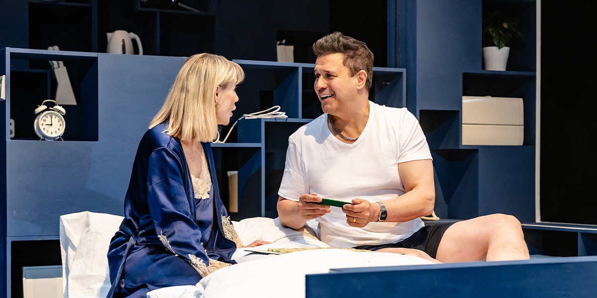 Two actors are on stage, seated in a bedroom setting, engaged in a conversation. The person o the right is holding a book and is laughing.