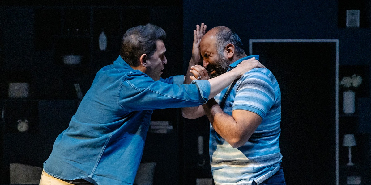 Two actors are on stage. The person on the right appears to be crying and the person on the left has both hands on the others shoulders. This is set against a backdrop that resembles a kitchen.