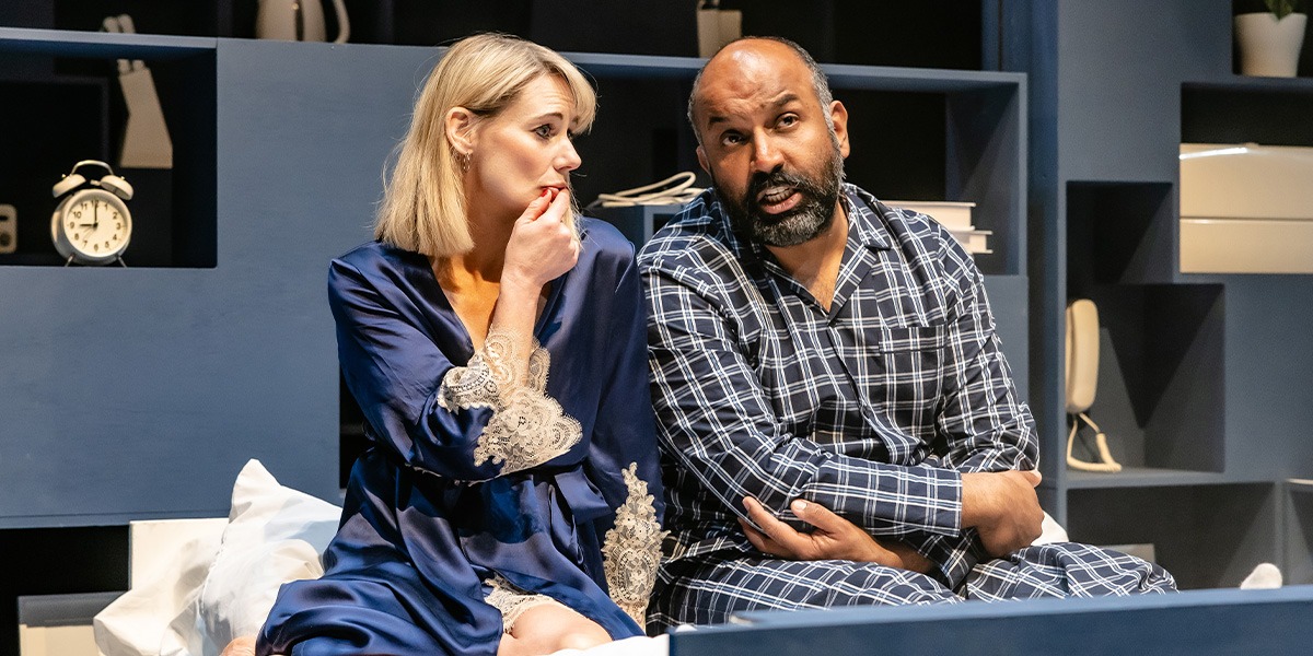 Two actors are on stage in a bedroom scene, both are dressed in bed clothes and appear engaged in a serious conversation.