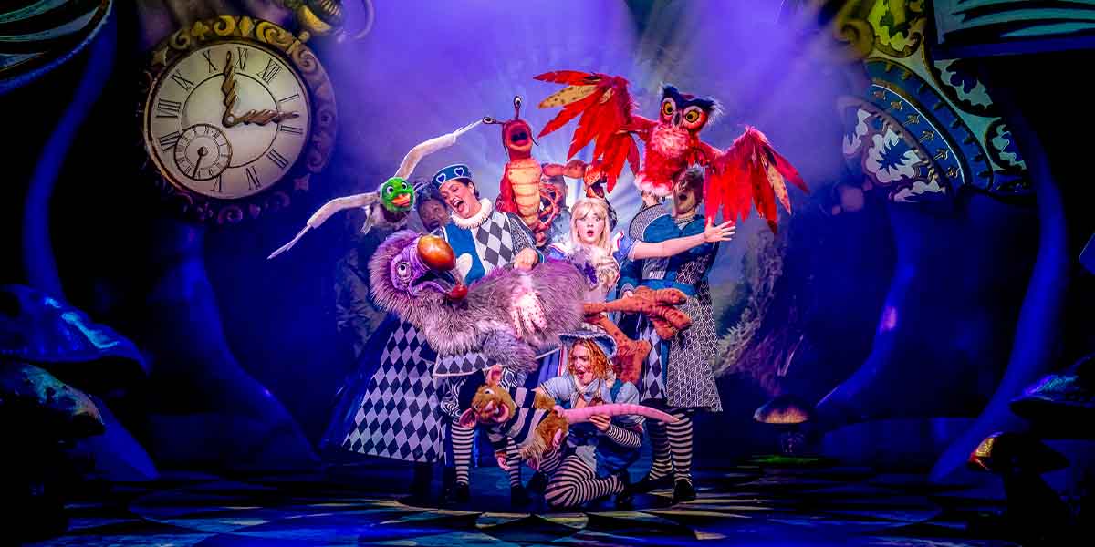 Charlotte Bradley as Alice poses with ensemble members, featuring vibrant costumes and whimsical puppets on stage.