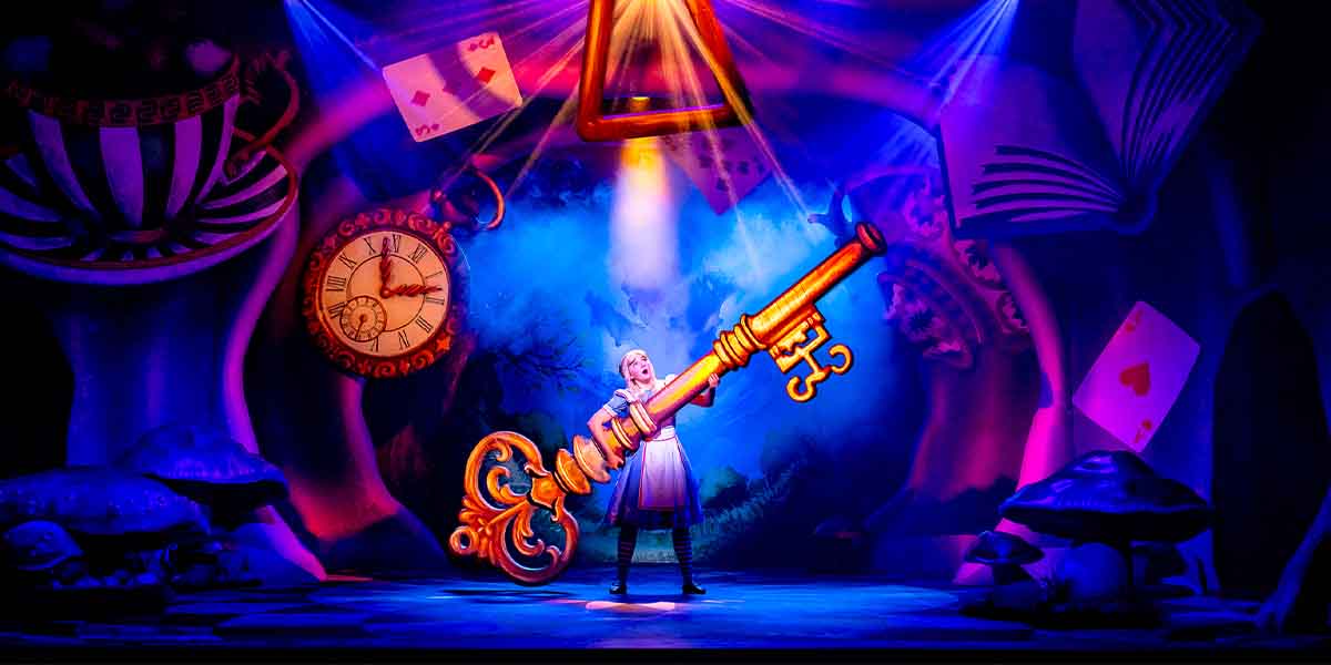 Charlotte Bradley as Alice holds a large golden key on a vibrant, whimsical stage backdrop.