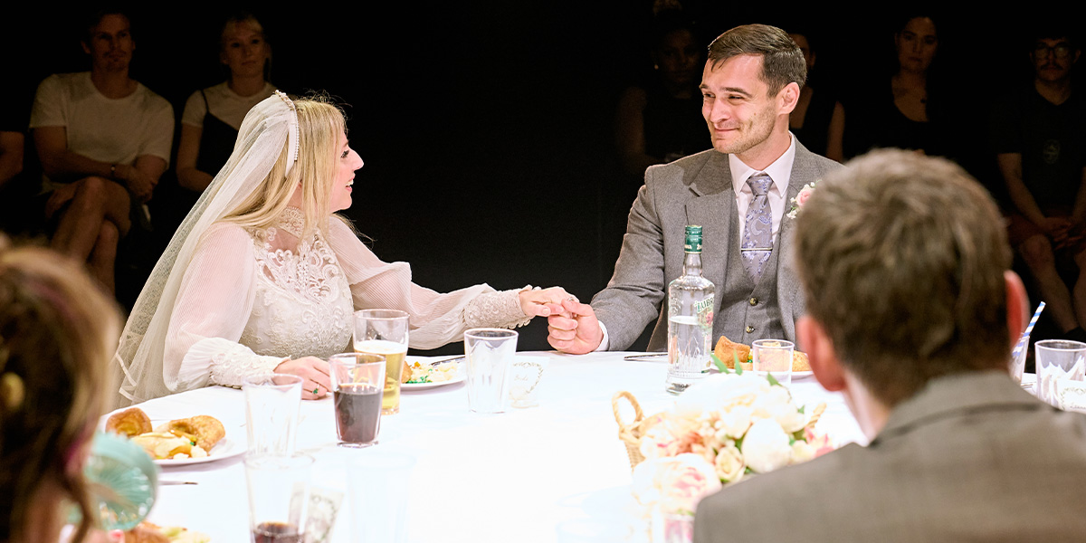 Two individuals in wedding attire are seated at a table, holding hands and looking at each other with a smile, during a wedding reception. There are drinks and food on the table, and guests can be seen seated around them.