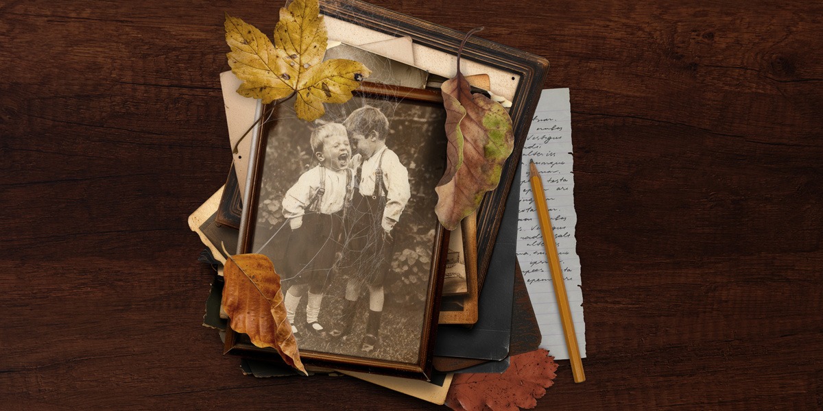 The background is a dark brown wood effect, like a table. In the middle of the image, is a framed, black and white photograph of two little boys. The one of the left is laughing, the one on the right has his head rested on the others head. They are both wearing white shirts with dungarees. The one on the left has white socks and black shoes, the one on the right has black socks and black shoes. They appear to be outside. There are leaves surrounding the frame. Behind them you can see bits of other frames of photographs, but not the photographs themselves. At the bottom of the pile of photographs, sticking out to the right, you can see a piece of paper with indistinguishable writing with a yellow pencil on top.
