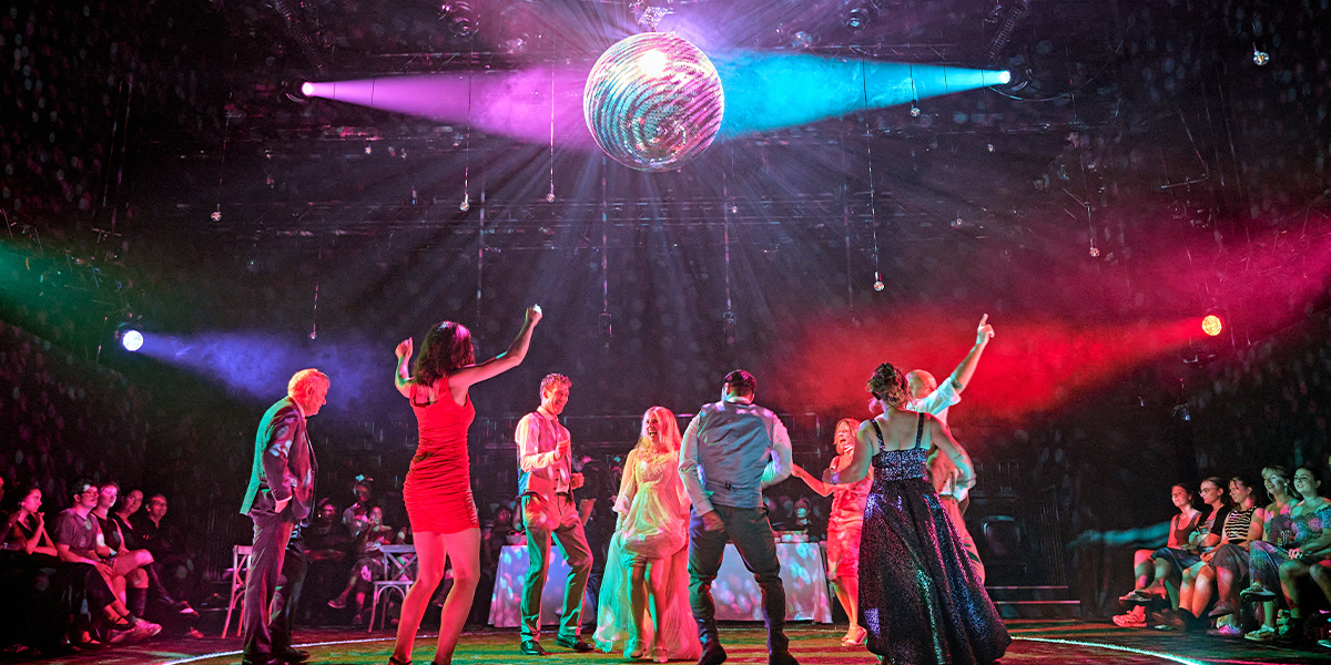 A vibrant scene from a musical performance, featuring a group of dancers under a large disco ball with colourful stage lighting.