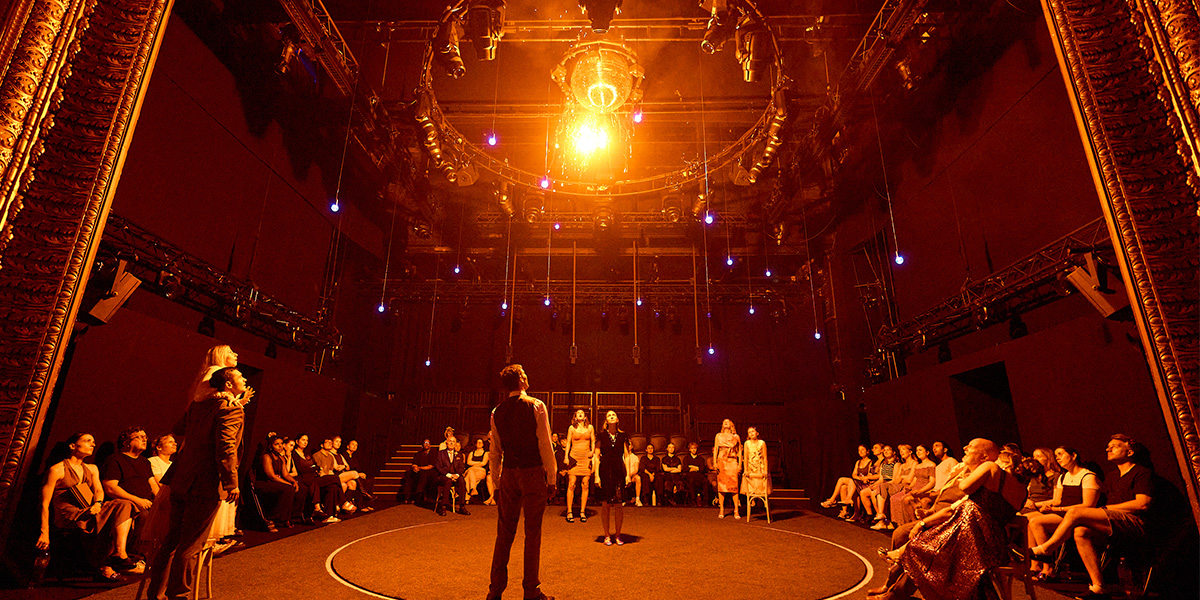 Audience members seated around a circular stage in a theater with warm ambient lighting and a central ornate chandelier, watching a performance.