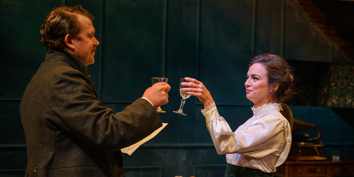 Two actors on stage in a theatrical production, toasting with champagne glasses in an elegantly designed set resembling a historical interior.